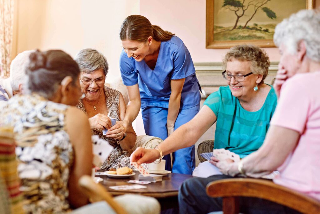 A caregiver spending time with a group of elderly women playing card games