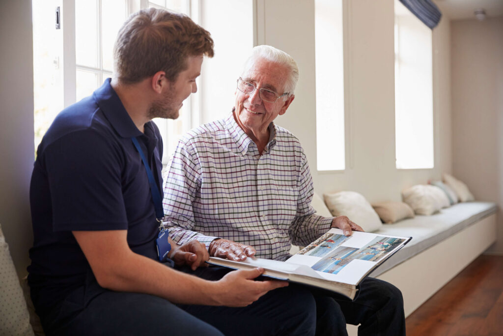 Senior man sitting looking at photo album with male caregiver