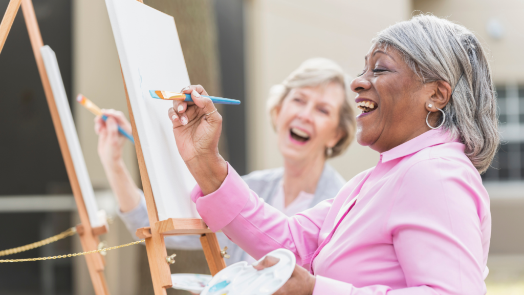 Two older women laugh and smile while painting on canvases outside